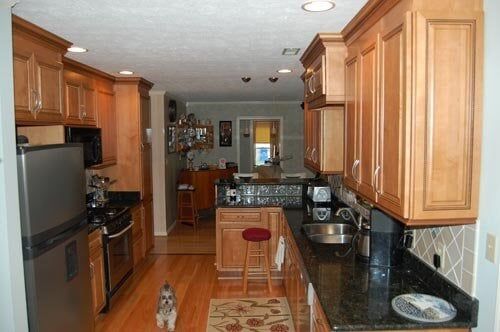 A galley-style kitchen featuring wood cabinets, dark countertops, stainless steel appliances, and a small dog on the floor.