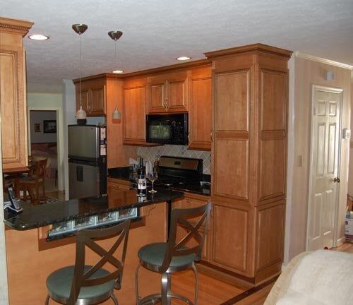 A small kitchen featuring natural wood cabinets, black countertops, stainless steel appliances, and two bar stools.