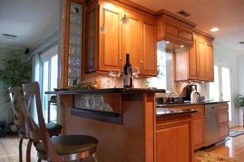 A kitchen island with two stools, light wood cabinets, and a wine bottle on the counter in a modern, sunlit home.
