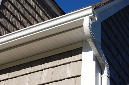 White house gutters and downspout attached to the corner of a building with beige cedar shake siding.