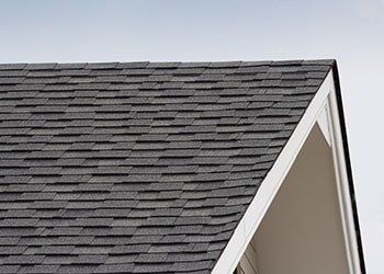 A close-up view of a dark gray asphalt shingle roof on a house with white trim against a plain sky.