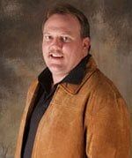 Herb Paynter—General Contractor in Newport News, VA A person with a slight smile wears a brown suede jacket over a black collared shirt against a mottled brown background.
