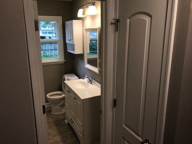 A small, neutral-toned bathroom featuring a white vanity, a toilet, a wall-mounted cabinet, and an overhead vanity light.