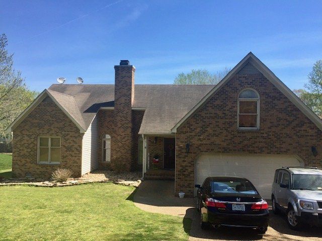 A two-story brick house with a brown shingled roof, a chimney, an attached garage, and two cars parked in the driveway.
