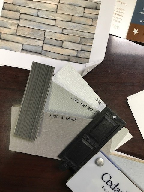 A fan of siding color samples in shades of gray, featuring a stone texture swatch, resting on a dark wood table.