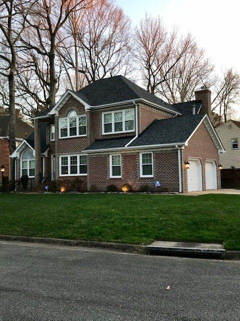 A two-story brick house with a dark shingled roof and an attached two-car garage, surrounded by grass and bare trees.