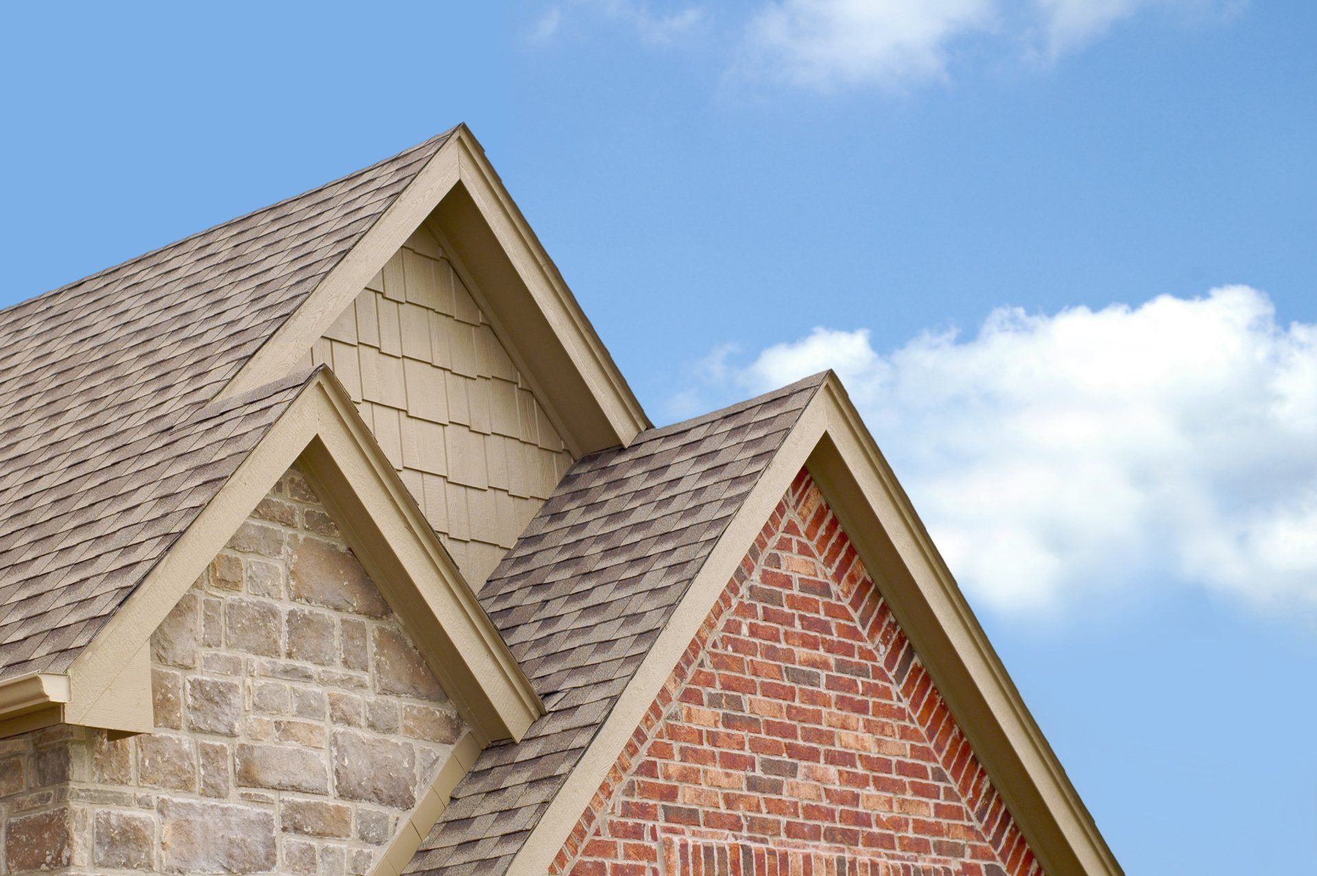 The peaks of a house roof featuring stone and brick siding against a blue sky with white clouds.