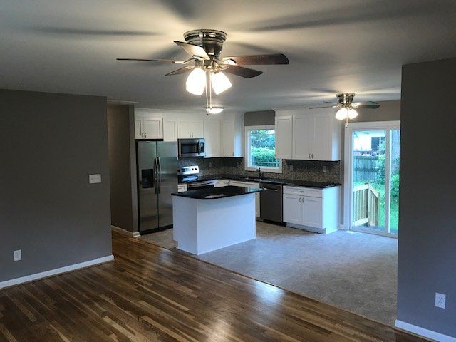 A wide-angle view of a kitchen with white cabinets, dark countertops, and a central island, opening to a carpeted area.