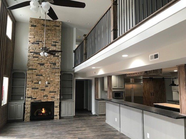 A living room with a tall stone fireplace, ceiling fan, and a loft overlook, transitioning into an open kitchen area.
