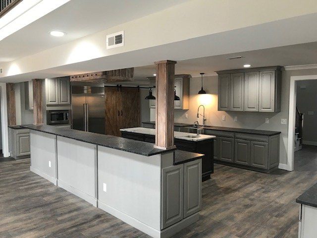 A modern basement kitchen featuring grey cabinetry, a long peninsula with black counters, and wood-paneled support columns.