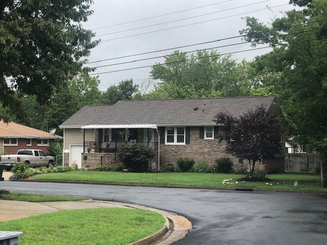 A tan, single-story house with a brick facade, a covered front porch, and a gray shingled roof on a suburban street.