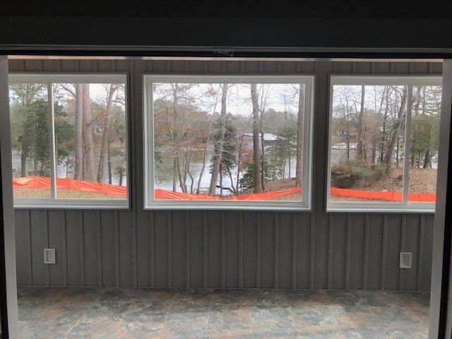 Three windows look out from a gray room onto a lake surrounded by trees with an orange safety fence in the foreground.