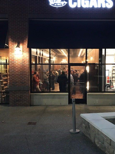 A group of people standing and talking inside a well-lit cigar shop seen through a large storefront window at night.