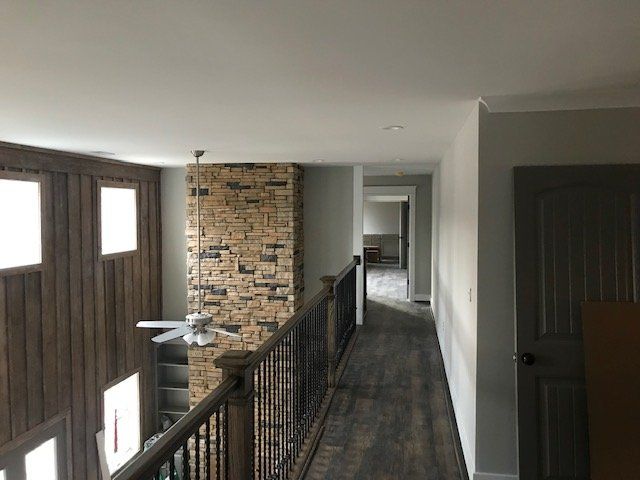 A hallway with dark wood flooring, a stone accent wall, and metal railings overlooking a room with a ceiling fan.