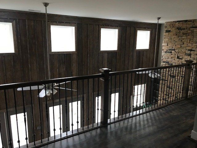 A wooden balcony with a dark railing overlooks a room featuring vertical wood-paneled walls, ceiling fans, and stone decor.