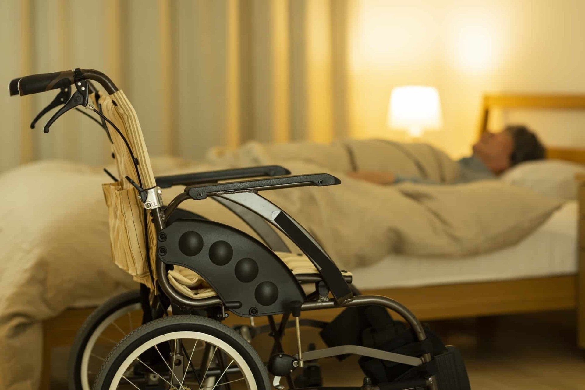A wheelchair sits in the foreground with a person sleeping in a bed in the softly lit background.