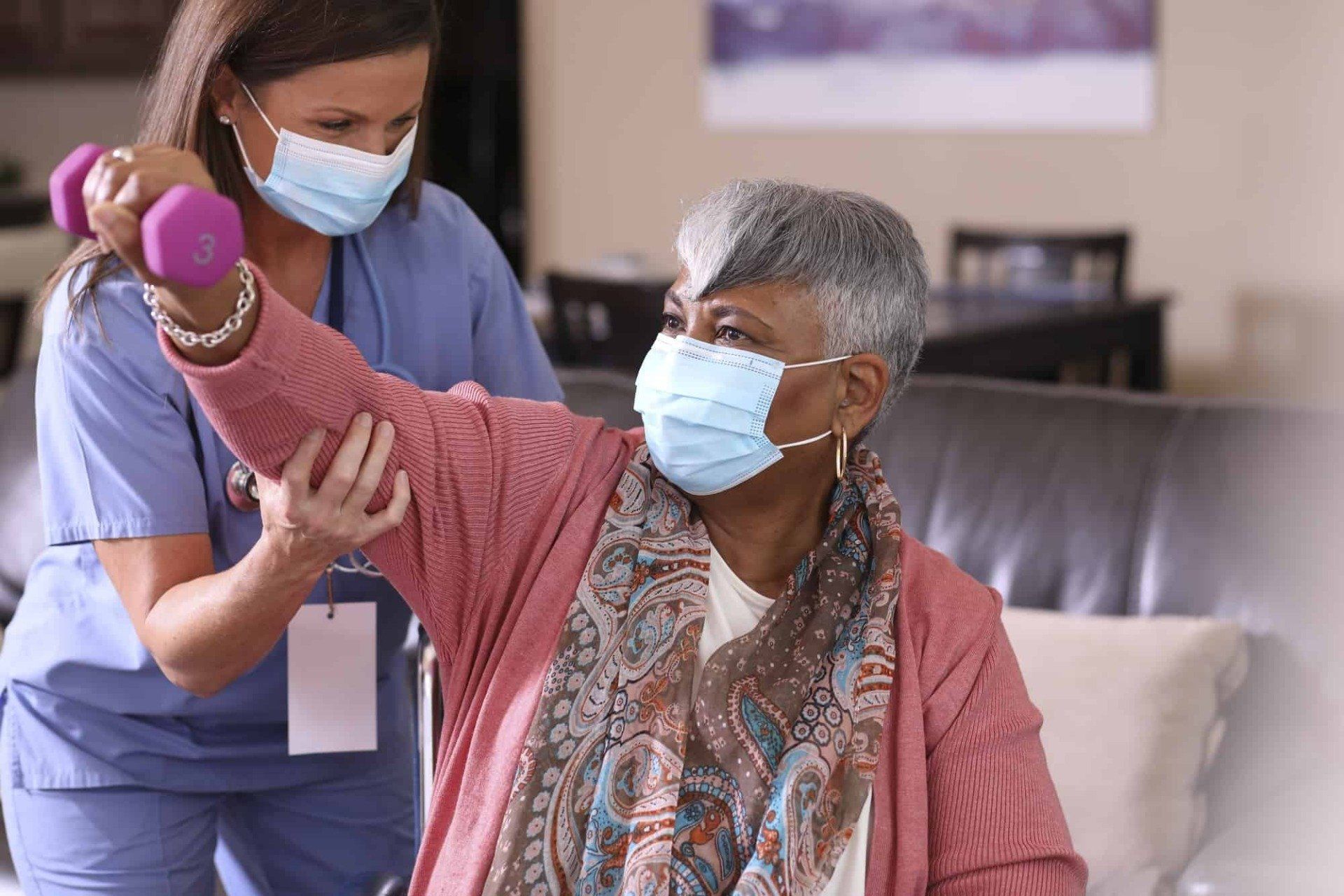 A healthcare worker in blue scrubs helps a person in a pink cardigan perform an arm exercise using a small purple dumbbell.