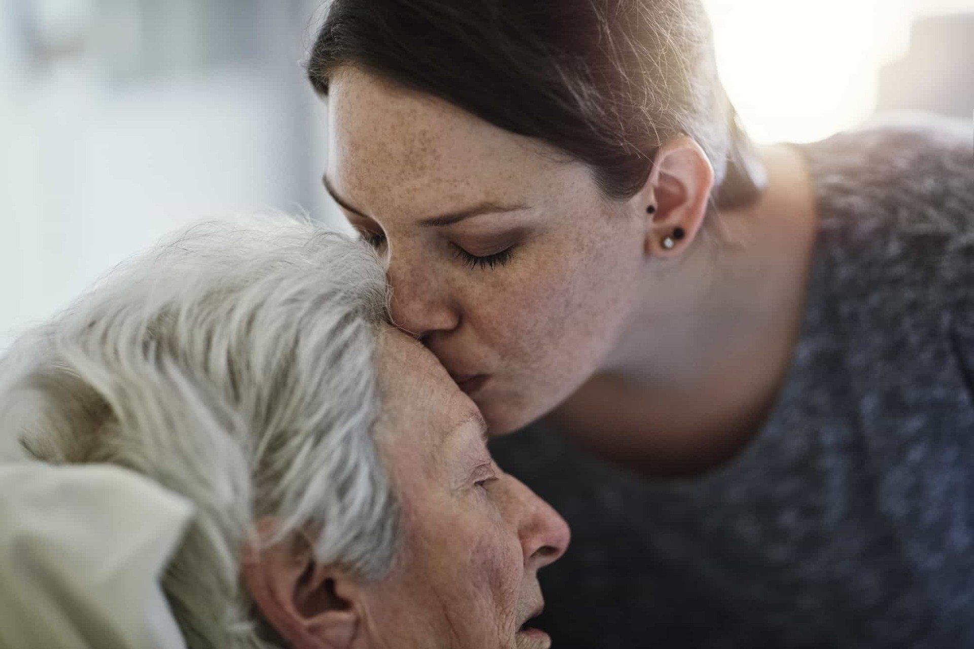 A person tenderly kissing the forehead of an individual with white hair who has their eyes closed.