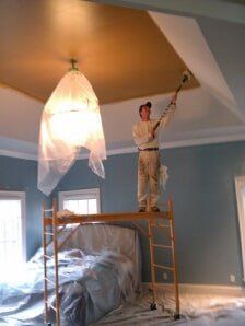 Person standing on a scaffold painting a bedroom ceiling around a hanging light fixture.