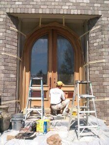 Worker kneeling on porch steps painting a wooden arched double door beside two ladders and paint supplies