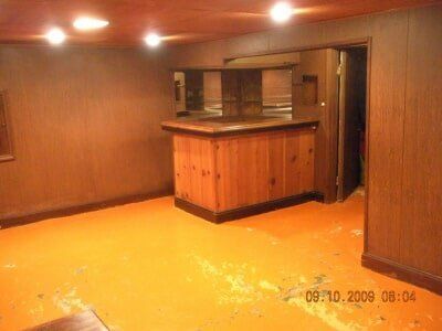 Empty wood-paneled basement room with orange floor and a small bar counter in the back