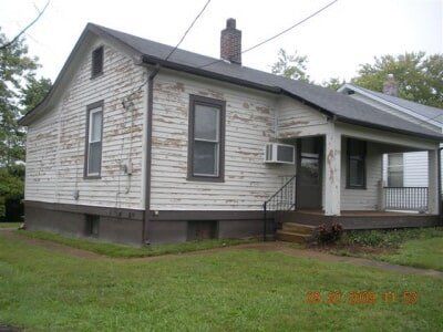 Small white house with a covered porch and dark trim, set on a grassy lot.