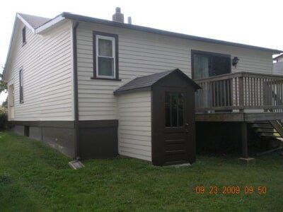 Small beige house with white trim, side deck, and dark brown shed in a grassy yard.