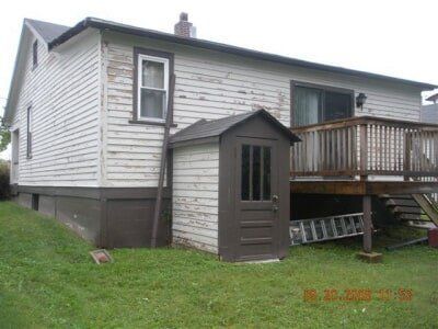 Gray house with white siding, small deck, and brown shed on a grassy hill