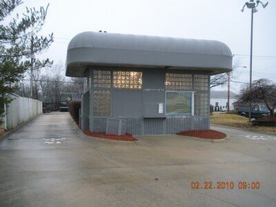 Small gray guard booth with a curved roof beside a wet driveway on an overcast day