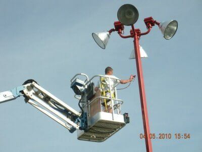 Worker in a lift servicing a red floodlight pole against the sky.