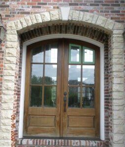 Wooden double front doors beneath a brick archway in a brick wall, with glass panes reflecting outside scenery