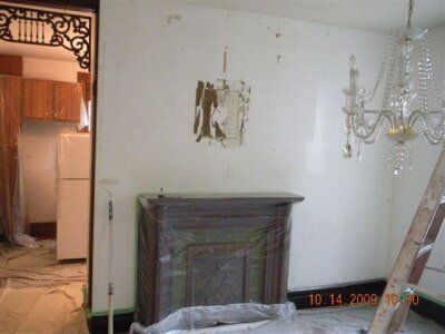 Damaged white room with bare walls, fireplace mantel, chandelier, and debris on the floor