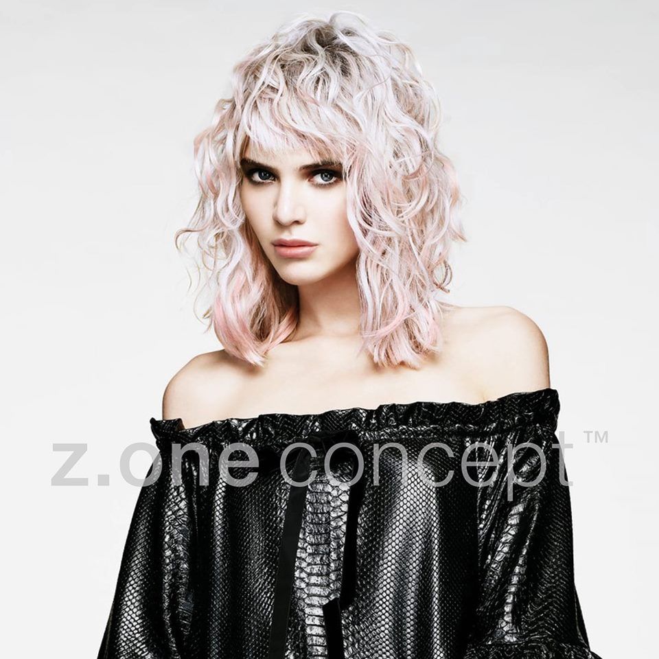 Mujer con cabello rosado y ondulado y top negro con hombros descubiertos, mirando directamente al espectador.