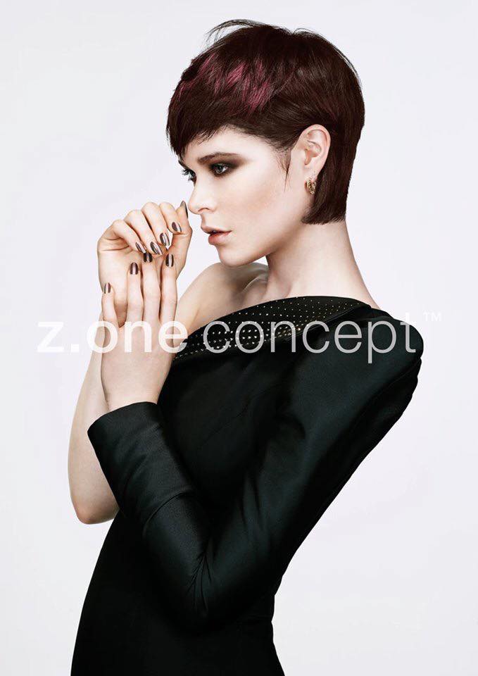Mujer con cabello corto de color rojo oscuro, con un vestido negro con hombros descubiertos, posando.