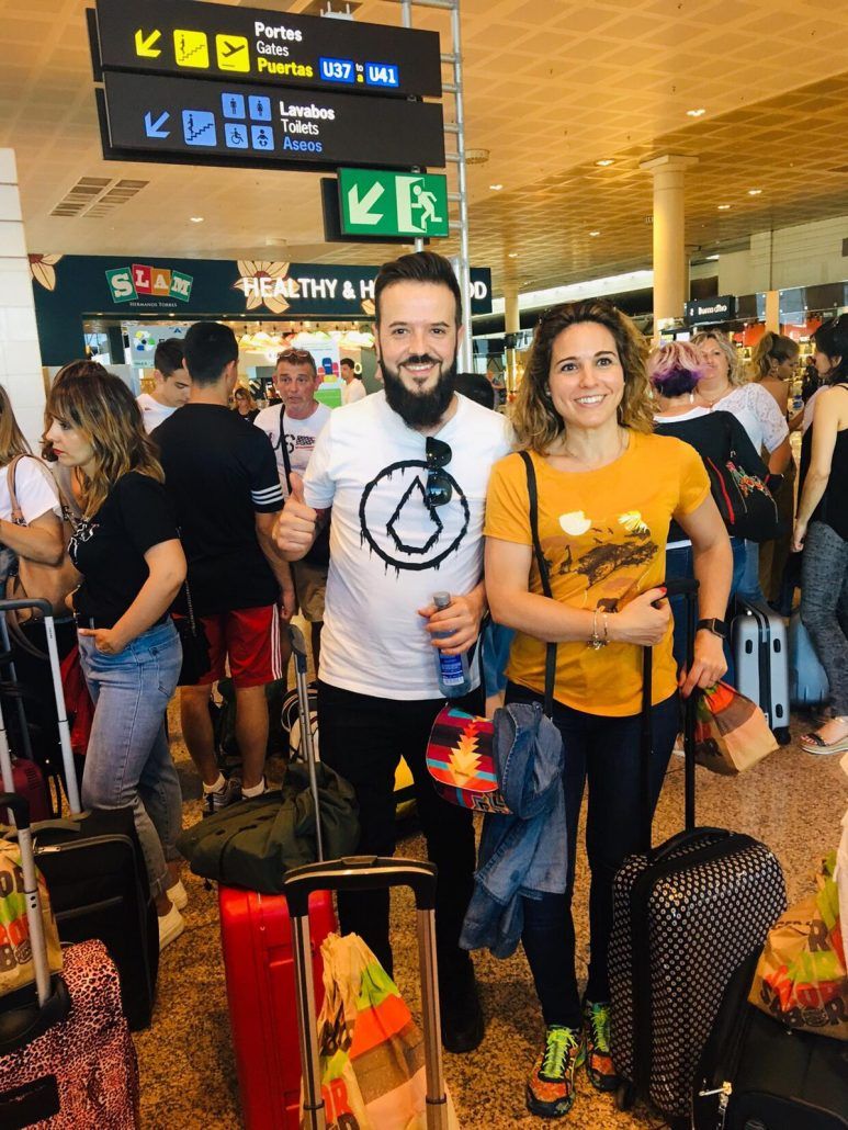 Hombre y mujer en el aeropuerto con equipaje, sonriendo y con el pulgar hacia arriba. Personas y carteles al fondo.
