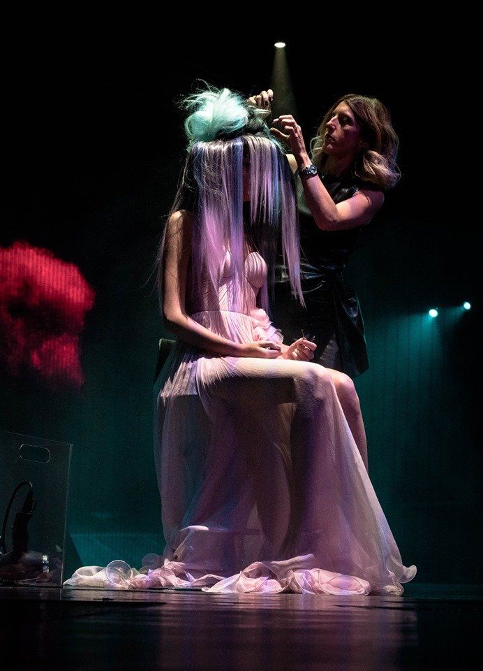 Mujer con vestido rosa peinándose en el escenario, entorno oscuro, bajo focos.