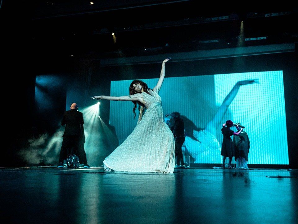 Una bailarina con vestido blanco posa en el escenario con el brazo extendido, bajo una luz azul. Otros artistas al fondo.