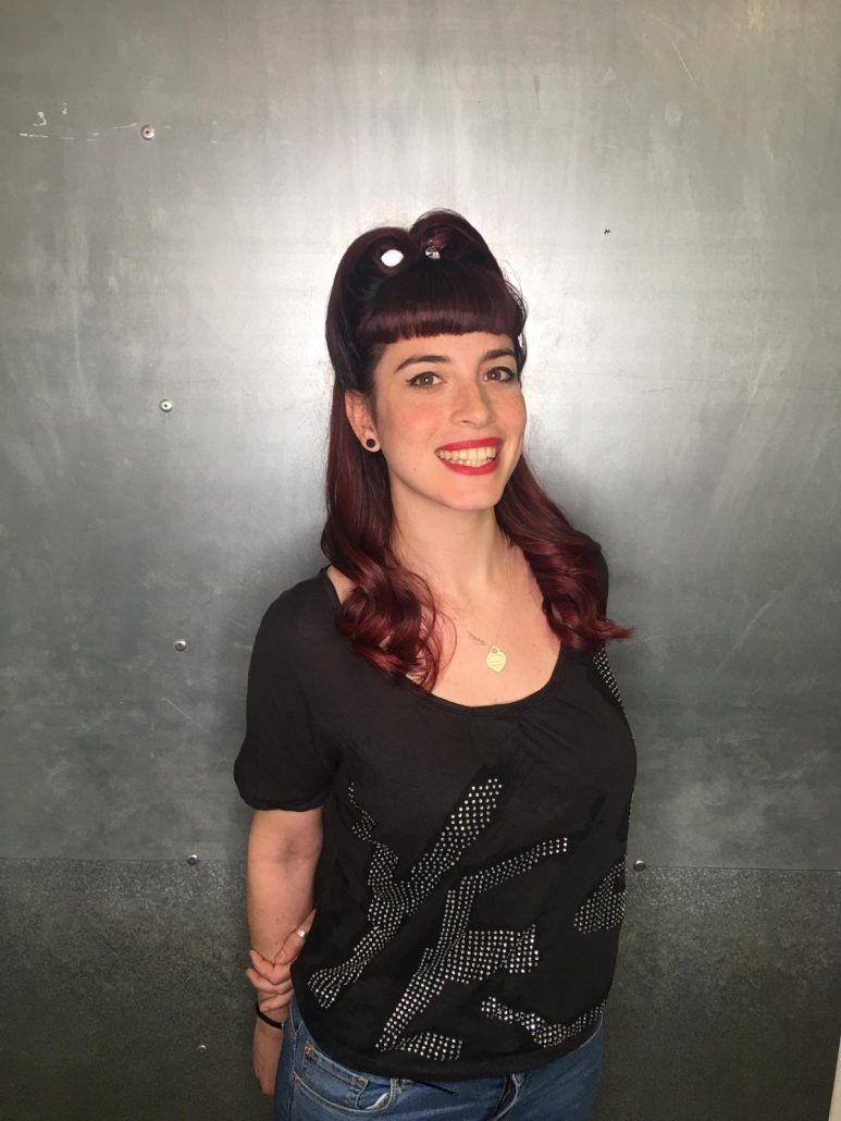 Mujer con peinado retro, cabello rojo y camisa negra sonríe frente a un fondo oscuro.
