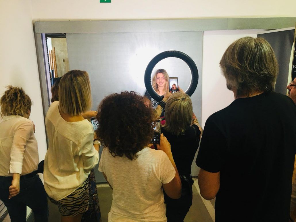 Una mujer se toma una selfie rodeada de personas que usan sus teléfonos en una habitación con un anillo de luz.