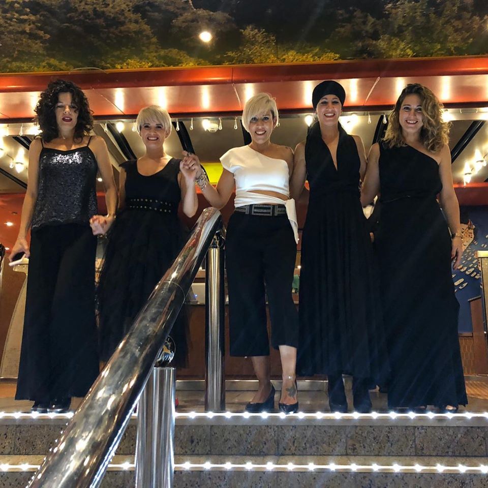 Cinco mujeres en las escaleras, sonrientes, vestidas de negro. Una con blusa blanca. Escenario nocturno con luces.