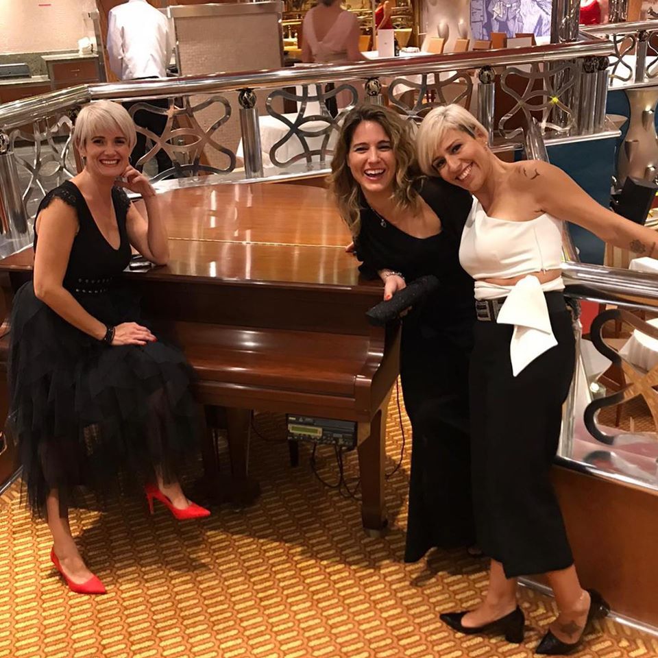 Tres mujeres sonrientes al piano. Una sentada, con vestido negro y zapatos rojos.