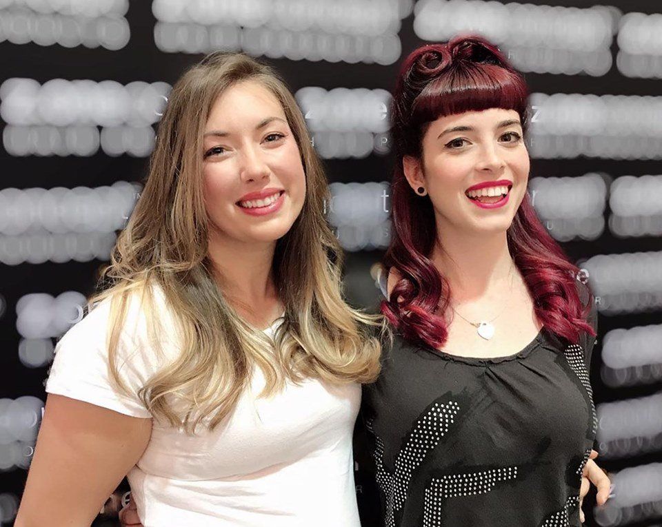 Dos mujeres sonrientes posando juntas. Una con camisa blanca, la otra con camisa negra y cabello rojo.
