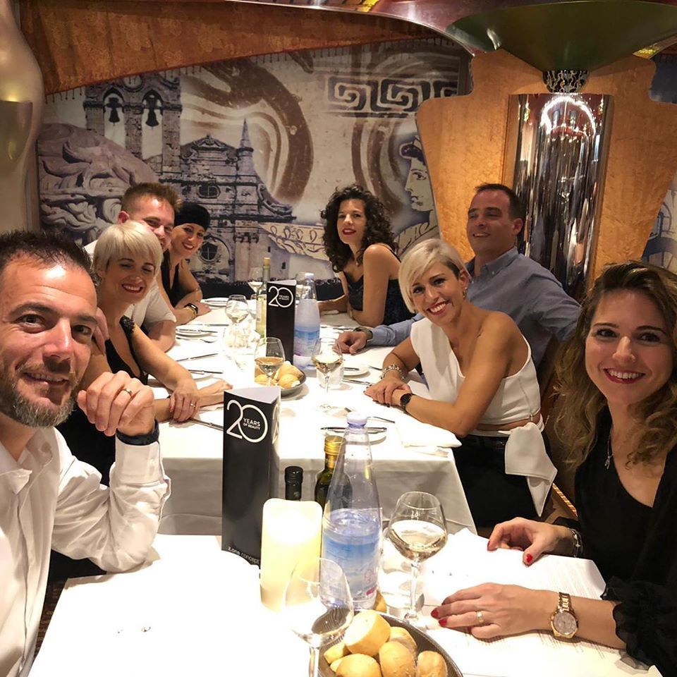 Grupo de personas en una mesa de restaurante, celebrando con bebidas. Expresiones alegres en una habitación con poca luz.