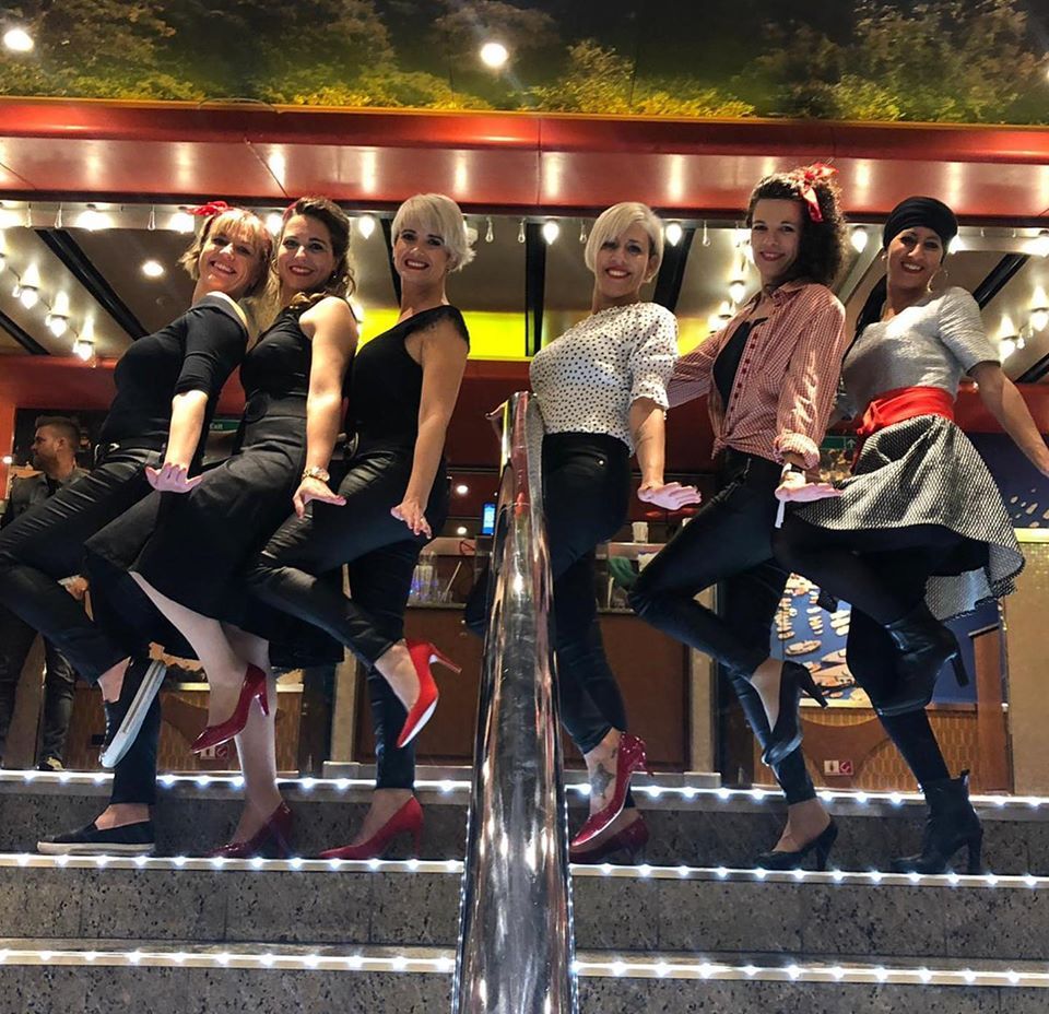 Seis mujeres con trajes de los años 50 posando en las escaleras; sonriendo, con detalles en rojo y negro.