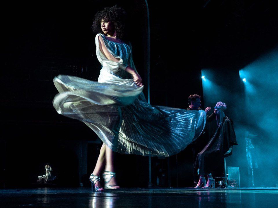 Una mujer con un vestido plateado baila en el escenario. Otros, sentados, observan bajo las luces.