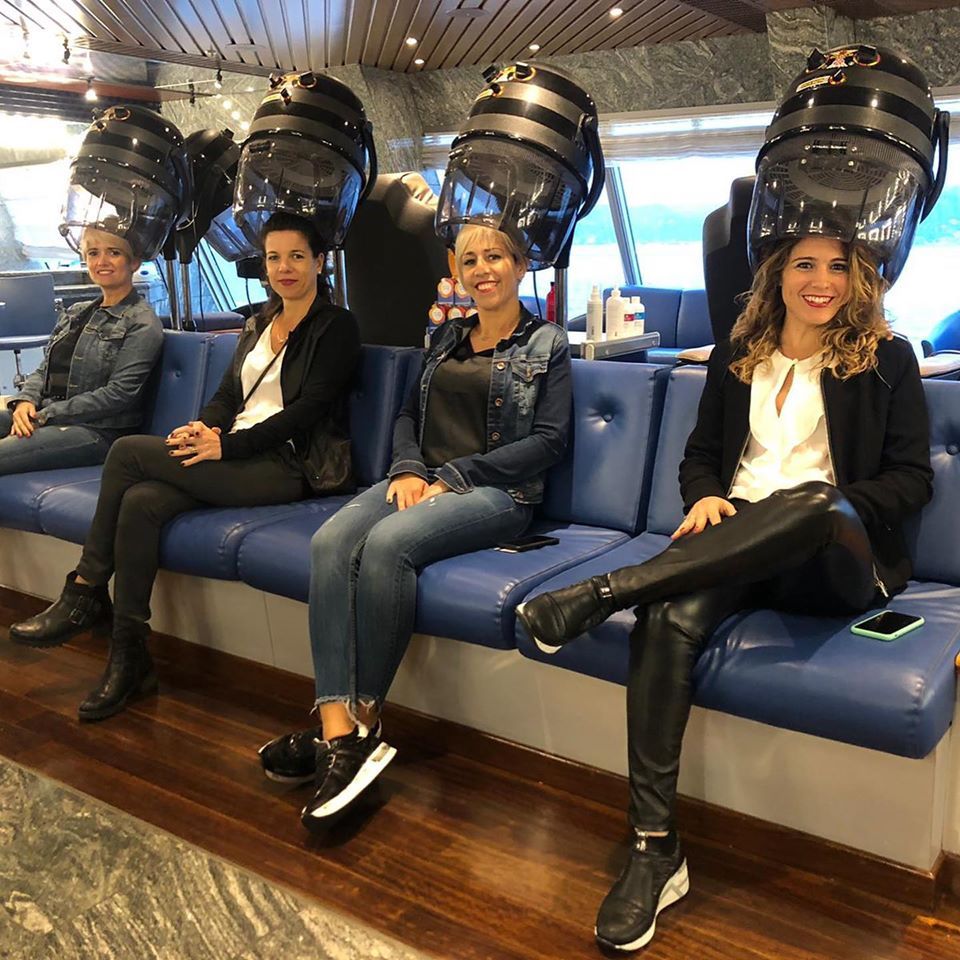 Cuatro mujeres sentadas bajo secadores de pelo en un salón; sonrientes, vestidas informalmente