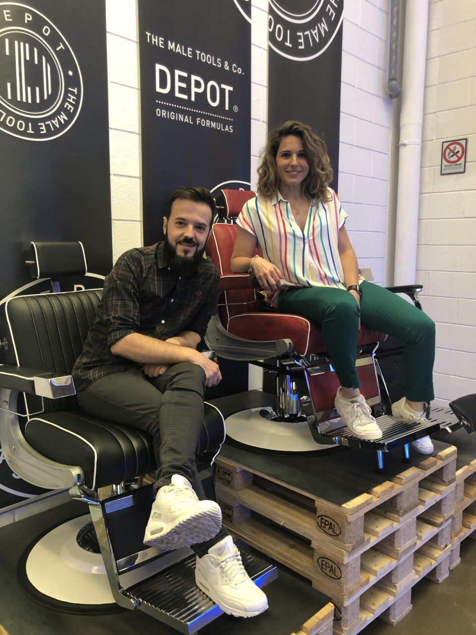 Dos personas, un hombre y una mujer, sentados en sillas de barbero con un fondo que dice