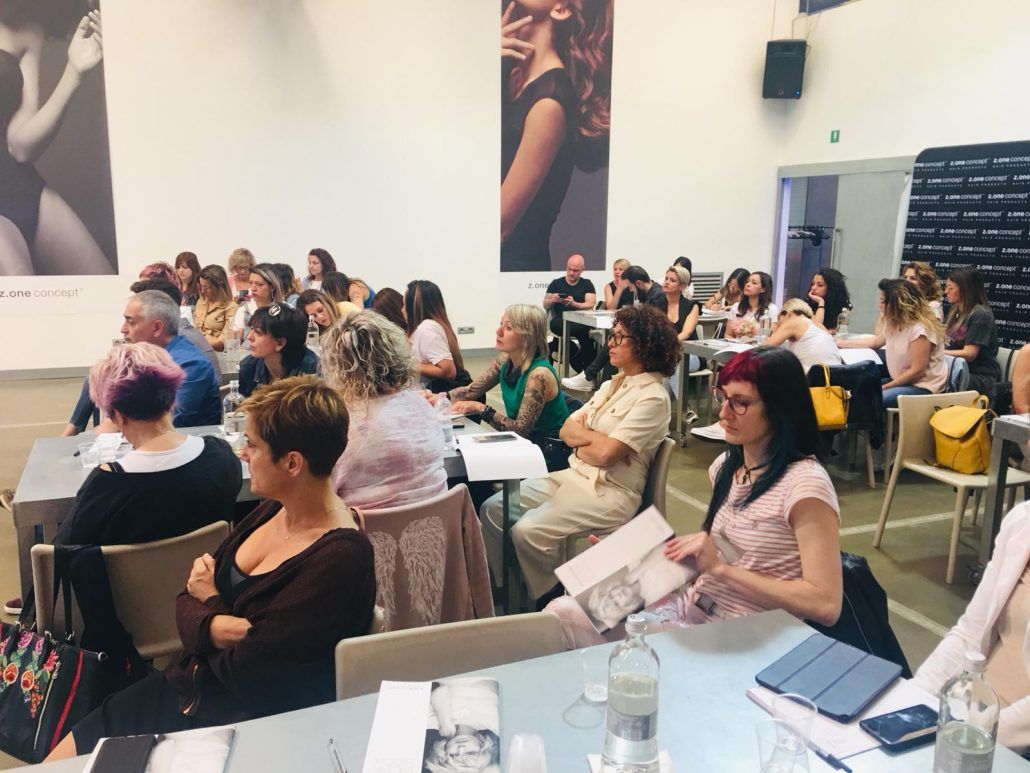 Público en una conferencia, sentados en mesas. Escuchando, algunos tomando notas. Grandes carteles en las paredes.