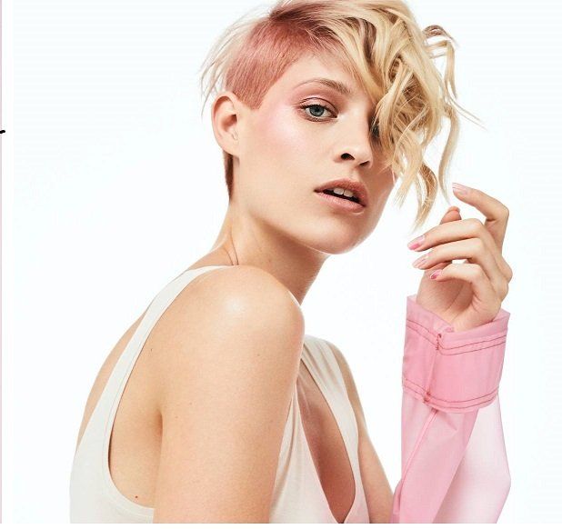 Mujer con cabello rosado y rubio mirando por encima del hombro, vistiendo un top blanco y una manga rosa.