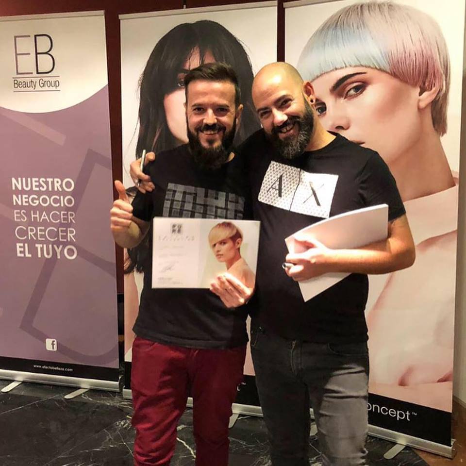 Dos hombres sonriendo, sosteniendo un certificado y papeles, en un evento de salón con pancartas.